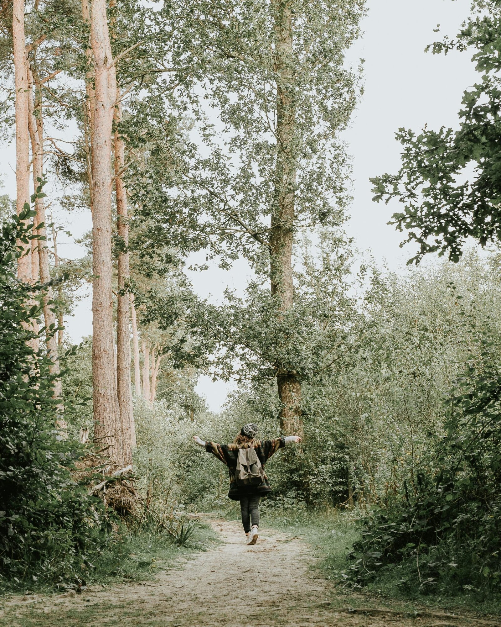 A woman joyfully walks down a serene forest pathway surrounded by tall green trees.